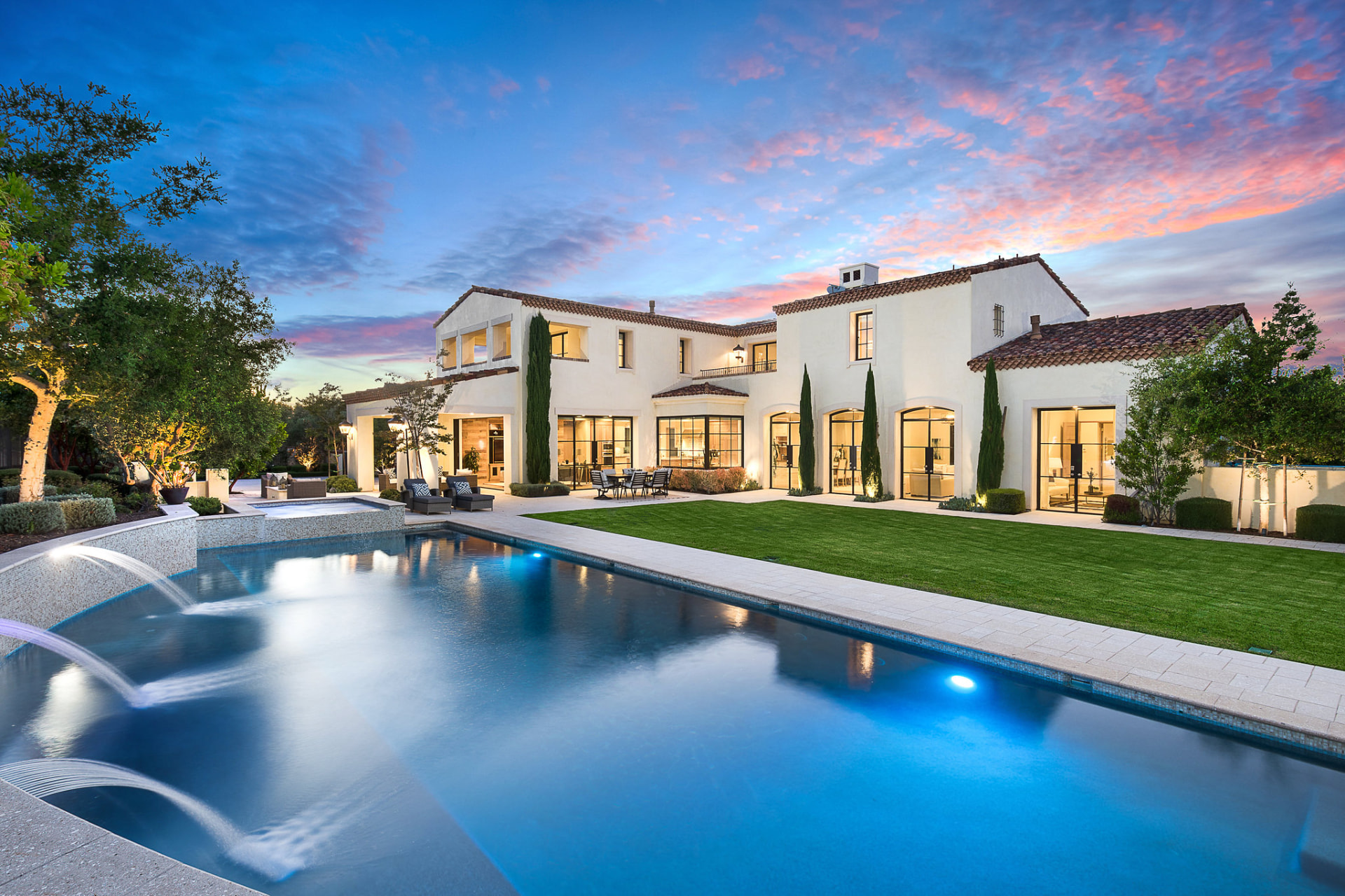 Mediterranean estate at twilight with a lit pool, fountain jets and pink evening sky