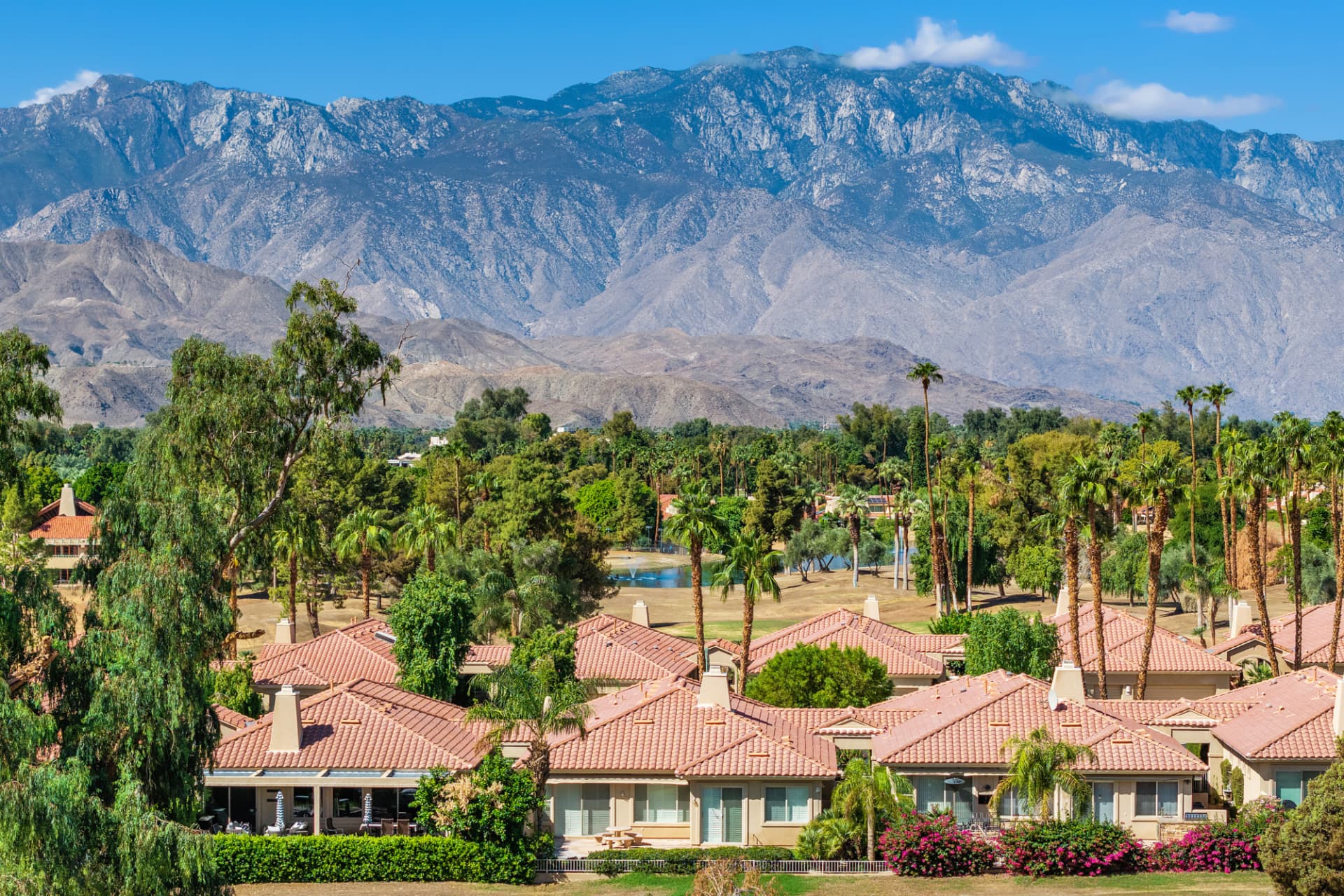 Aerial of a tile-roofed golf community with a fountain pond, palm trees and a rugged mountain range behind