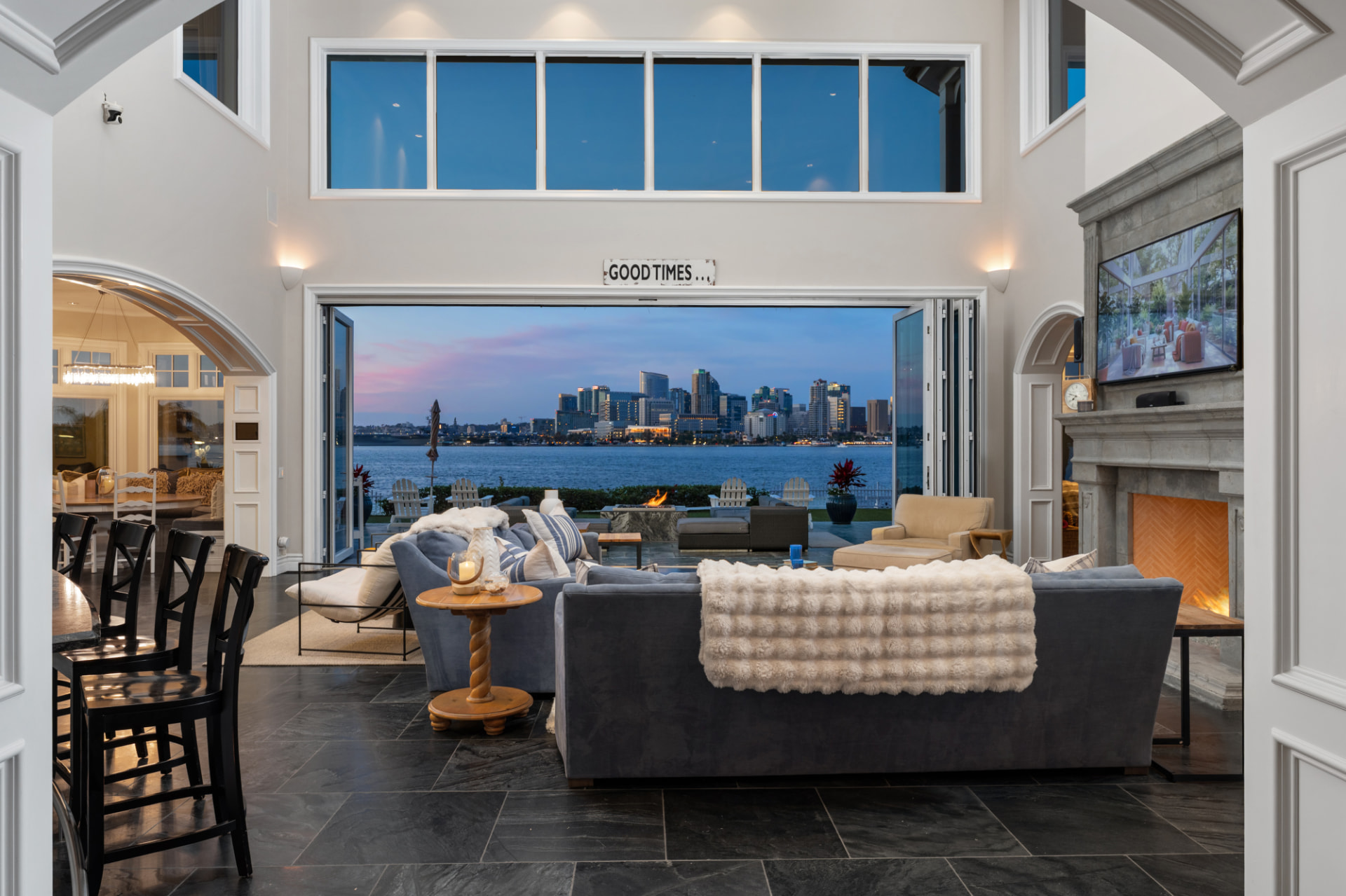 Waterfront living room with folding glass doors open to a patio fire pit and a city skyline across the bay at dusk