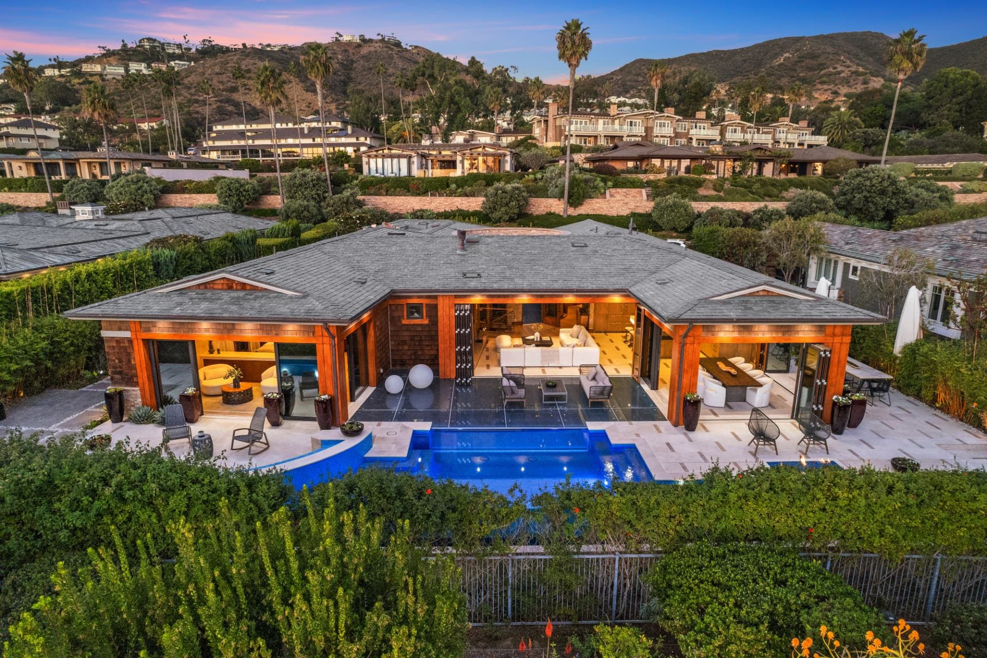 Twilight aerial of a single-story modern home with an open patio, lit pool and spa on a landscaped hillside